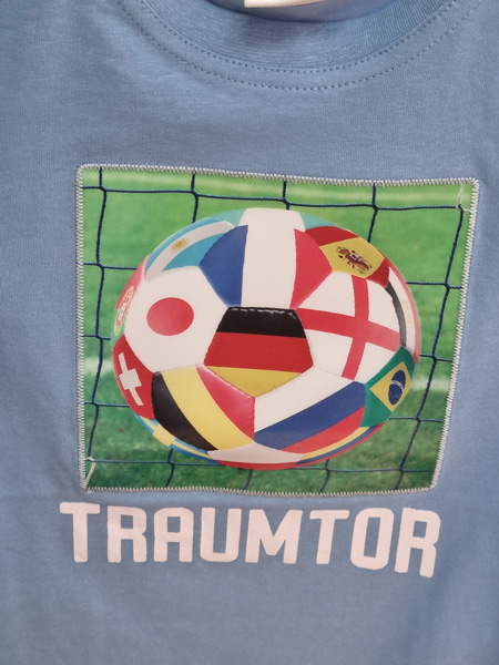 Blaues T-Shirt mit Fußball-Design, bedeckt mit Länderflaggen, auf grünem Feld. Text: "TRAUMTOR".