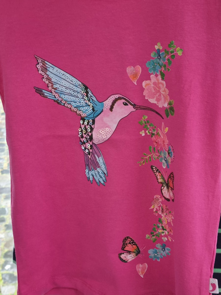 Ein pinkes T-Shirt zeigt einen bunten Kolibri mit Pailletten, umgeben von Blumen und Schmetterlingen.
