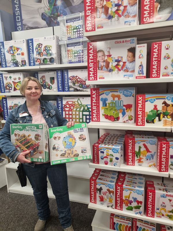 Eine Frau in Jeans hält lächelnd zwei bunte "Smartivity"-Spielzeugboxen in einem Spielzeuggeschäft.